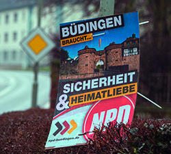 Abgebildet ist ein Wahlkampfplakat der Nationaldemokratischen Partei Deutschlands. Darauf steht: Büdingen braucht Sicherheit und Heimatliebe. Es ist eine Ansicht des Büdinger mittelalterlichen Untertors, auch Jerusalemer Tor oder Kreuztor genannt, abgebildet. Auf der unteren Bildhälfte befinden sich die Internetadresse der Büdinger Nationaldemokratische Partei Deutschlands und das Logo der Partei.