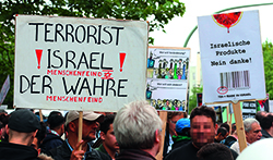 Abgebildet sind Demonstranten, die während des al-Quds-Tags in Berlin an Holzstangen befestigte Plakate tragen. Auf einem Plakat steht: Terrorist Israel! Menschenfeind! Der wahre Menschenfeind. Auf einem anderen Plakat steht: Israelische Produkte nein danke!
