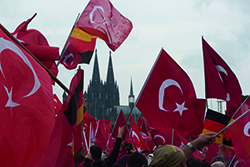 Abgebildet sind zahlreiche großflächige türkische Fahnen bei einer Demonstration in Köln, in kleinerem Format befinden sich auch deutsche Fahnen darunter. Dabei ist in der Ferne der Kölner Dom zu sehen.