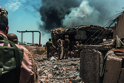 Abgebildet sind ein ehemaliges Kampfgebiet des Islamischen Staats im Irak, massive Kriegszerstörungen und aufsteigender Rauch, der von den Zerstörungen rührt.