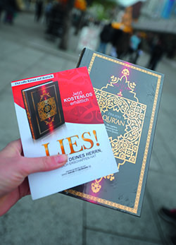 Abgebildet ist die linke Hand einer Person, die in einer Fußgängerzone eine Ausgabe des Koran und einen Flyer des LIES-Projekts hält.