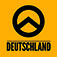 Abgebildet ist das Logo der Identitären Bewegung Deutschland. Auf einem rechteckigen gelb-orangenem Hintergrund befindet sich in schwarzer Farbe ein Kreis, darin der griechische Buchstabe Lambda, also ein Winkel, etwas größer als 90 Grad, dessen Spitze nach oben zeigt. Die beiden Enden des Winkels gehen in den schwarzen Kreis über. Darunter steht in schwarzen Großbuchstaben das Wort Deutschland. 
